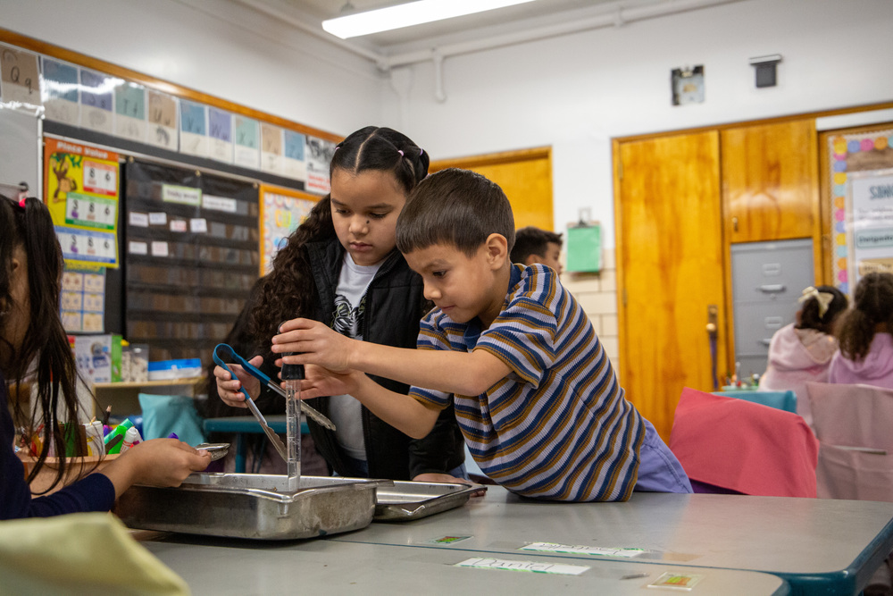 Two students attempt to clean debris from water using a baster and tongs. 