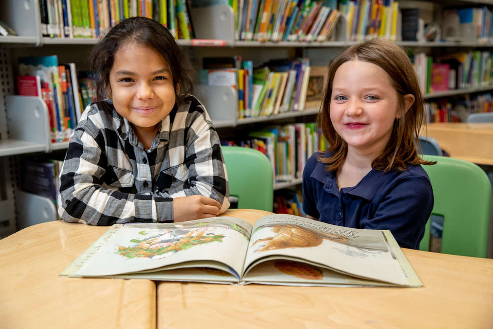 Two elementary students sitting at table  with book and smiling at camera