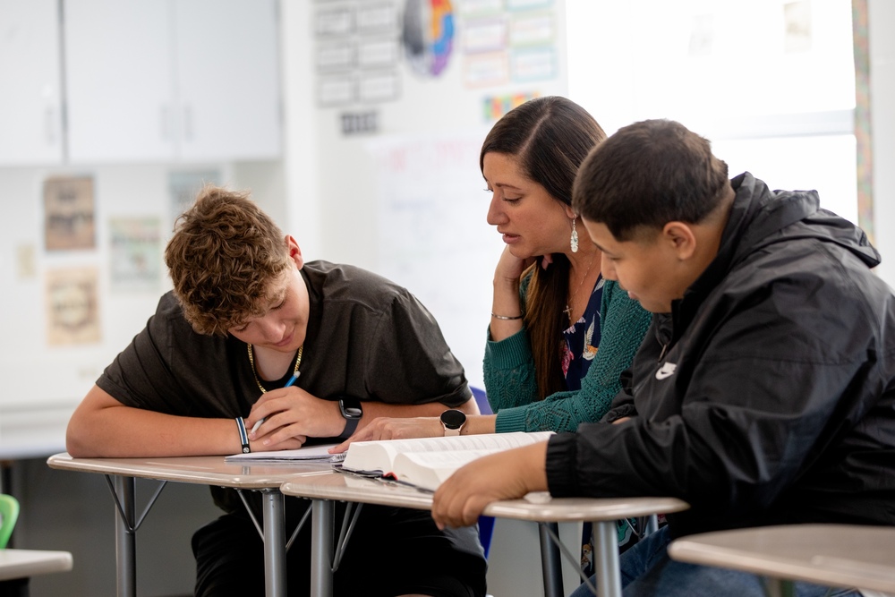 two students at their desks with their teacher reading a book