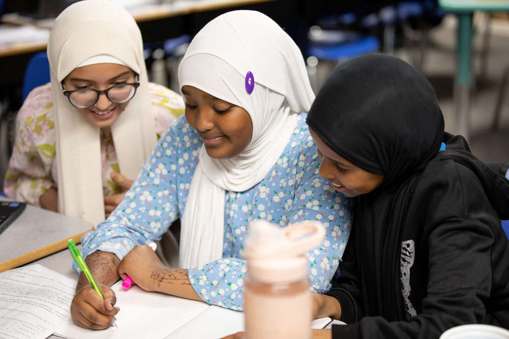 Three secondary students sitting at desk smiling at workbook