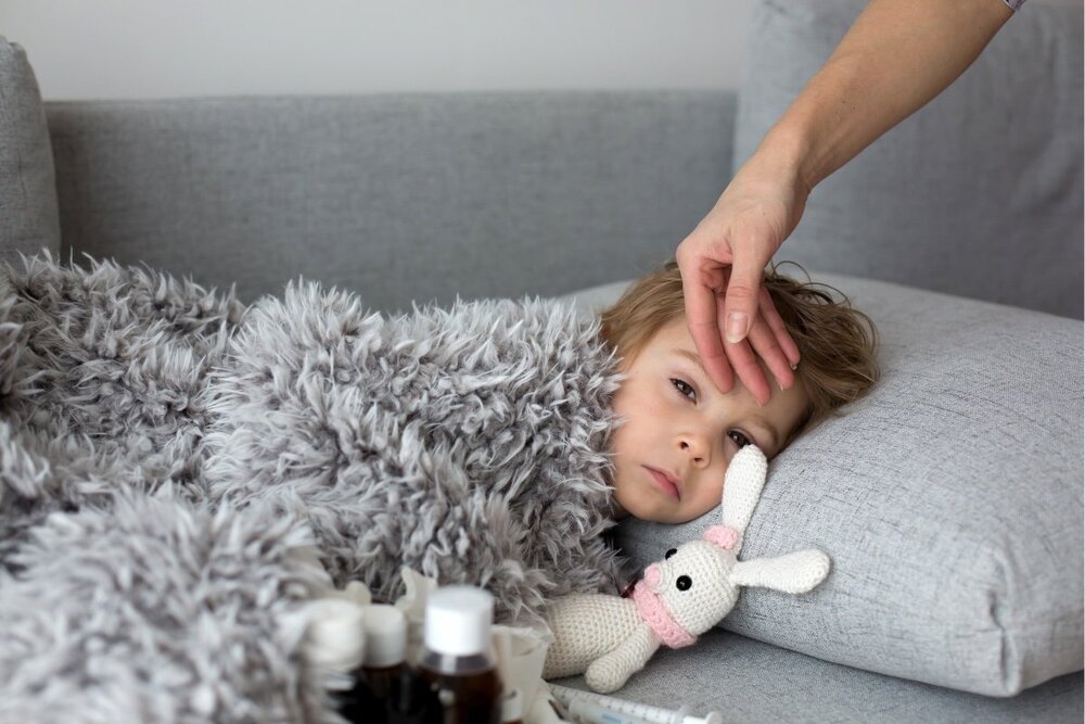 a sick toddler lays on a couch with a crochet bunny rabbit as a woman's hand feels  her forehead to check for fever