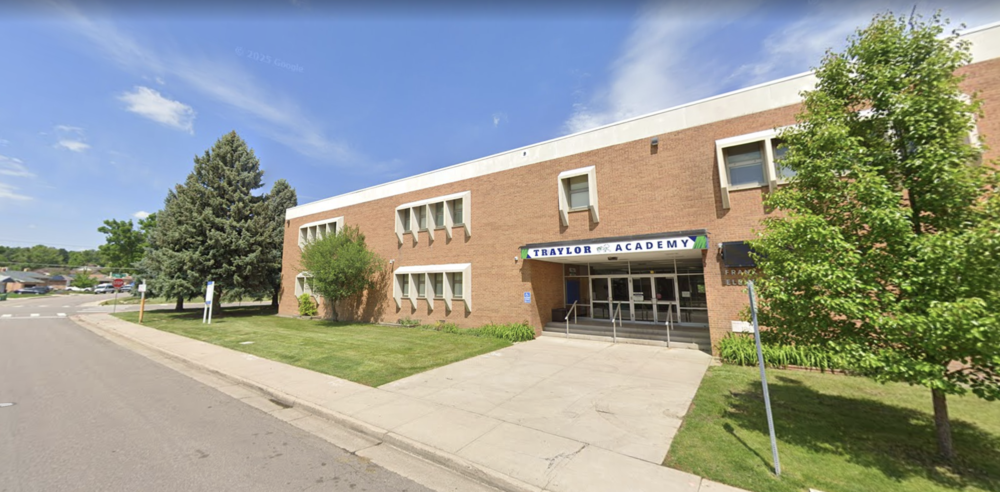 **Texto alternativo en español:** “Edificio de ladrillo de Traylor Academy, con un letrero sobre la entrada principal, rodeado de césped, árboles y una calle residencial bajo un cielo azul despejado.”
