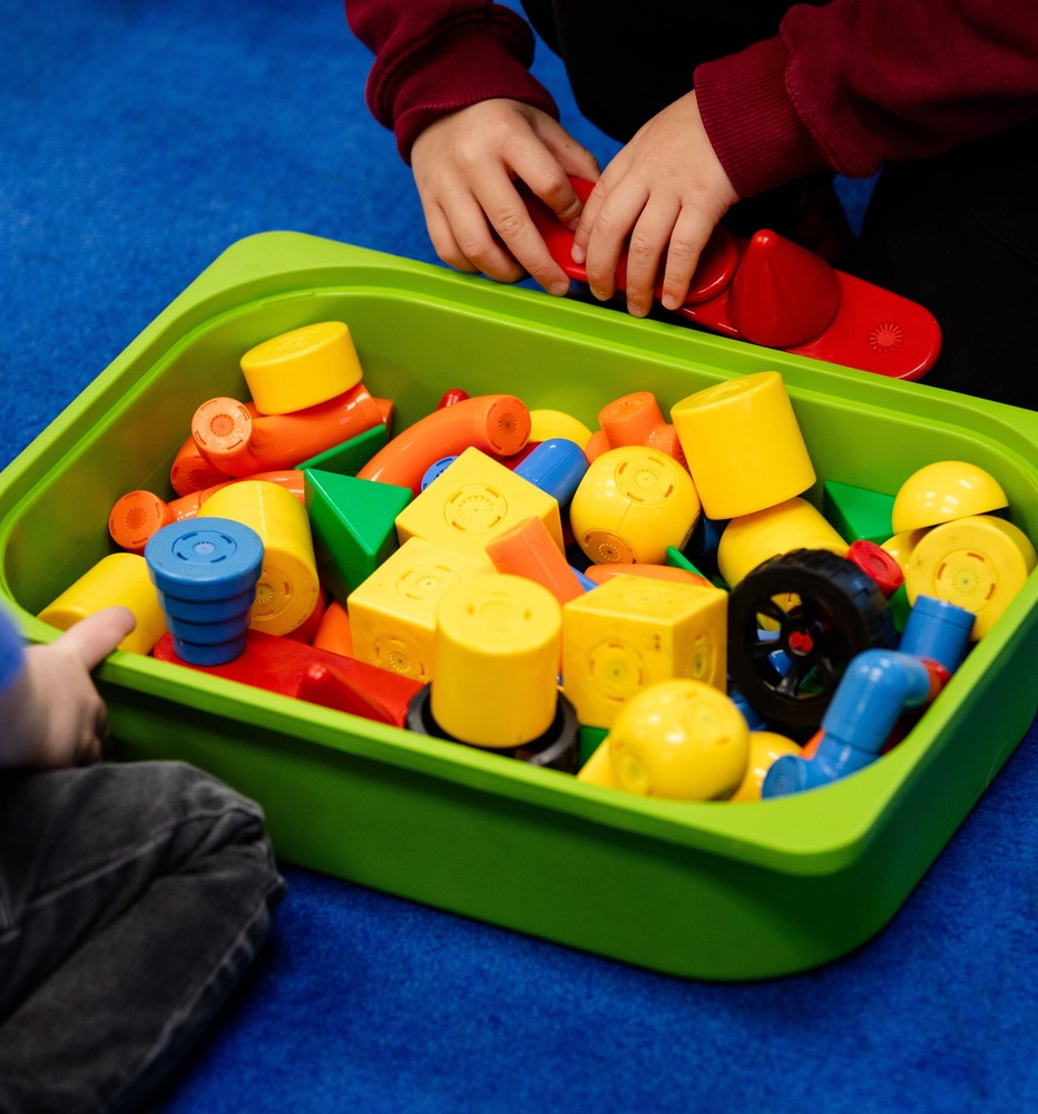 two children reach for multicolored blocks in a green bin