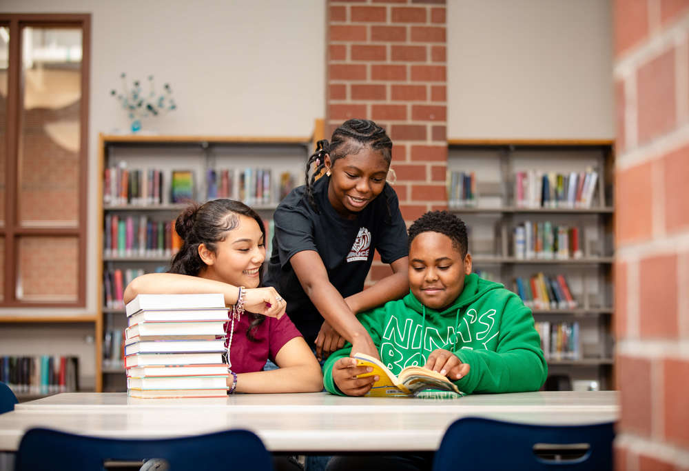 hree students sitting at a table with a stack of books in the library