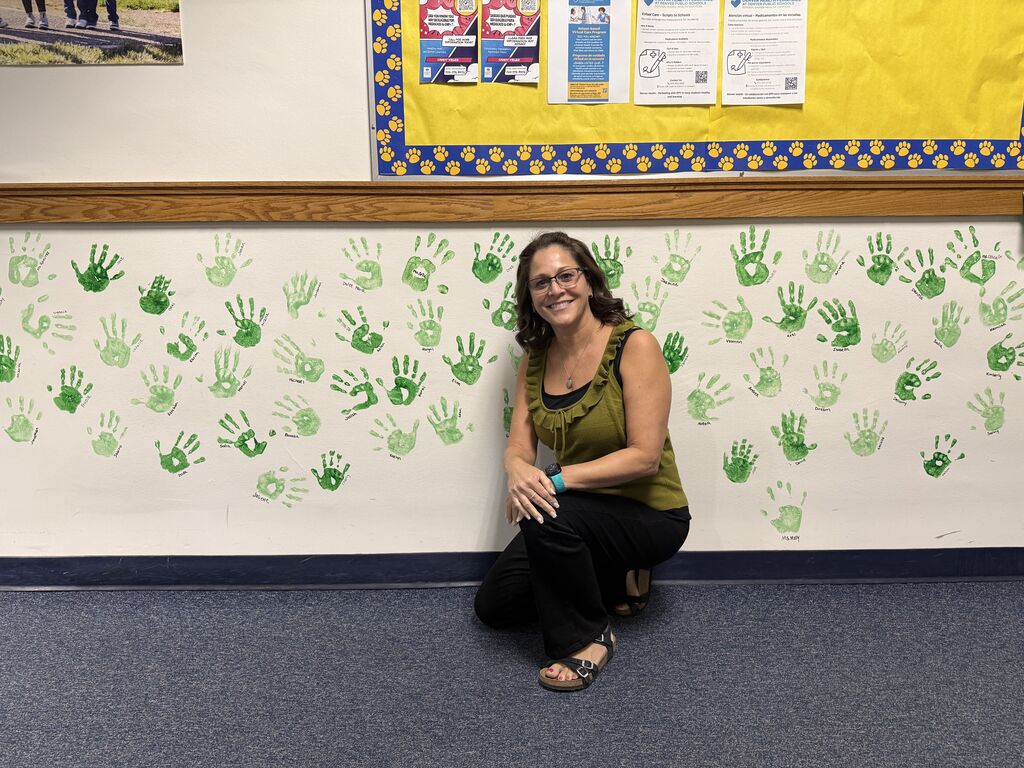 Principal kneeling in front of a white wall with green handprints. Papers are pinned to the upper wall.