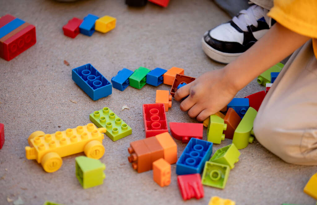 A child wearing a yellow shirt, black and white shoes plays with scattered colorful building blocks on a gray floor.