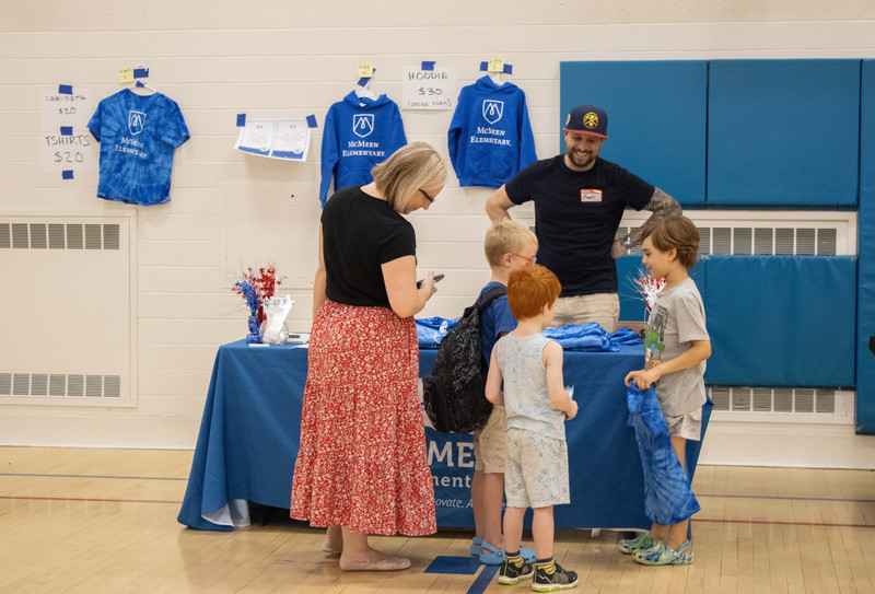 McMeen students chatting with a staff member at a table in the gym. McMeen swag and gear hangs on the wall behind them.