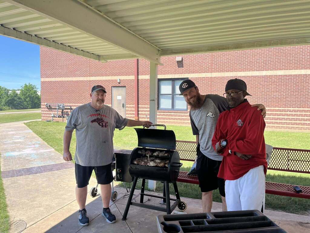 Coach Boren, Coach Carter, and Mr. Johnny stadning next to a smoker cooking chicken thighs