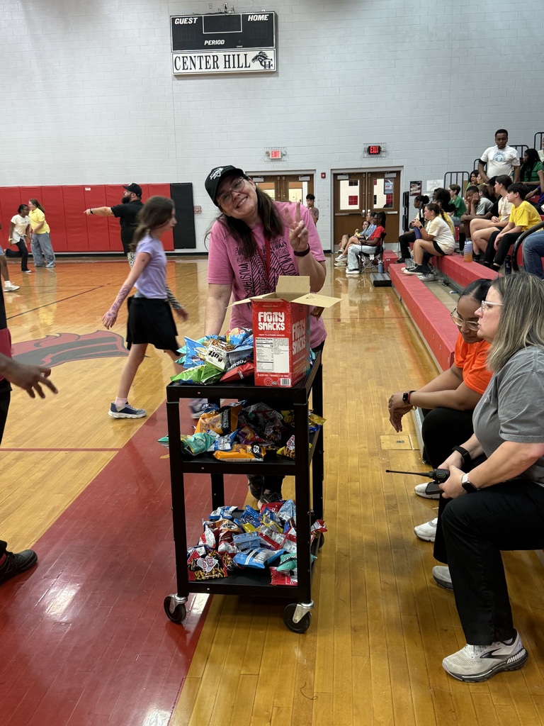 Teacher in the gym pushing a two tiered snack cart full of bags of chips,  cookies, and fruit snacks in the gym  with two teachers looking at the snacks .