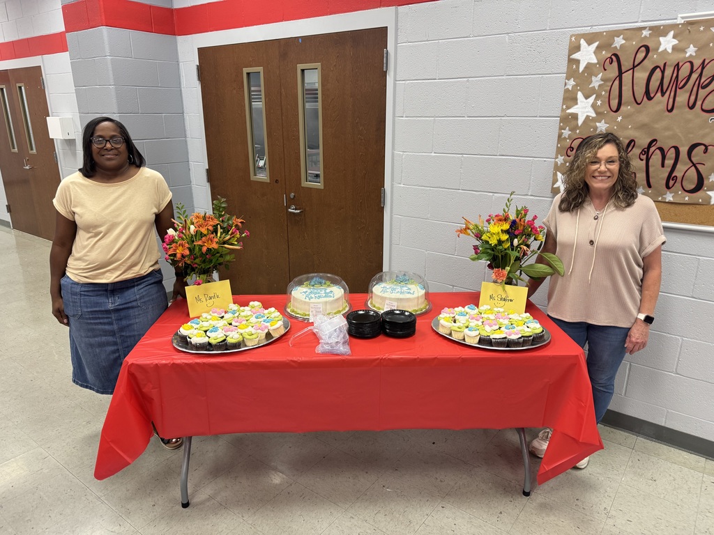 Two adult women standing on either side of a table. The table has a red table cloth, two small cakes, and two trays of cup cakes.  There are two vases with orange flowers and two cards. One card says "Mrs. Randle" and the other card says "Mrs. Shoffner. 