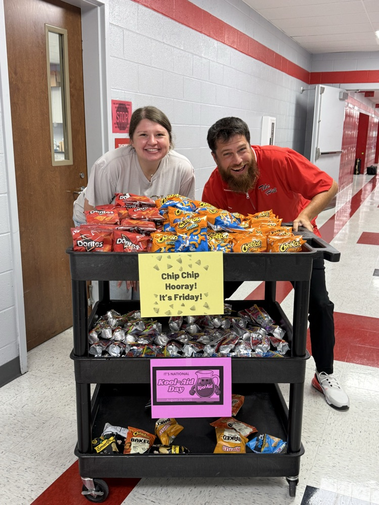 Two teachers smiling behind a snack cart filled with a variety of bags of chips and a variety of Kool-Aid Jammers.