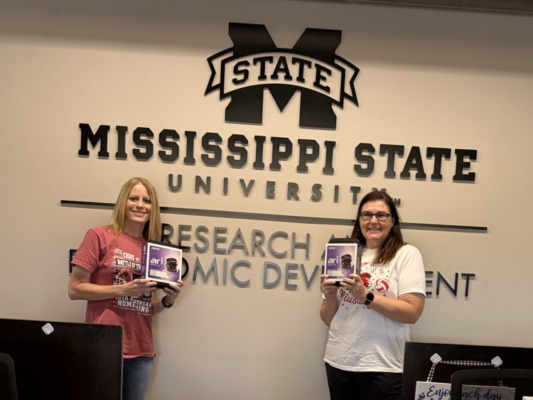Image shows Mrs. Hileman and Mrs. Palmer holding Ozobots in front of a MS State sign.