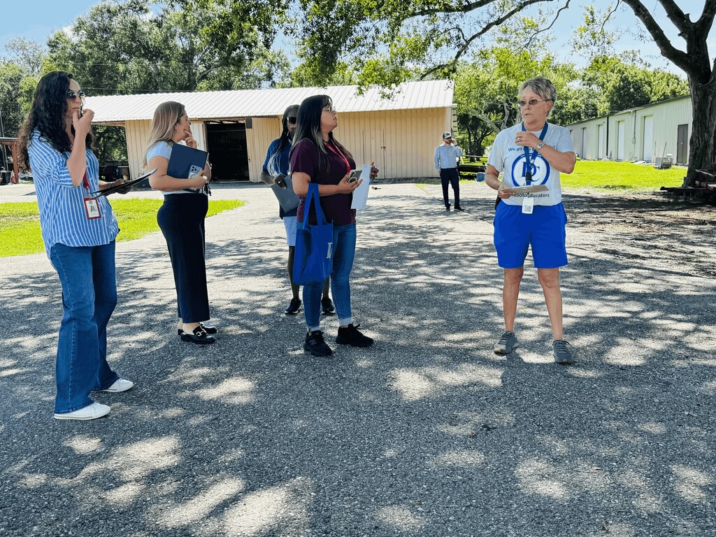 woman wearing grey and blue, holding a clip board and  talk ing to a group of women