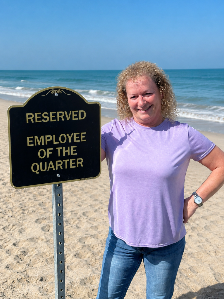 woman standing on a beach wearing jeans and a purple shirt with a sign that says "reserved employee of the quarter"
