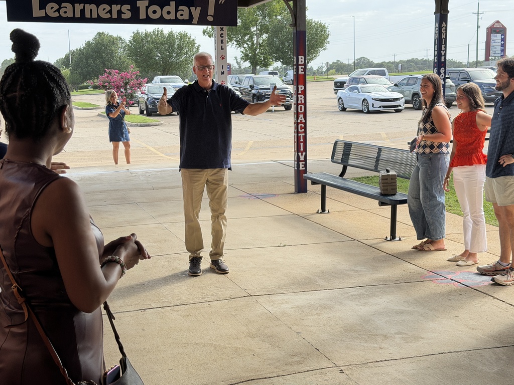 A man speaks with a group of people on a platform. A sign reads "Learners Today!" A bench is behind them.