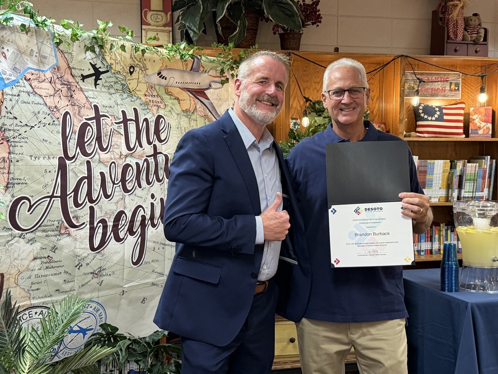 Two men stand in front of a decorated wall and bookshelf. One holds a certificate.