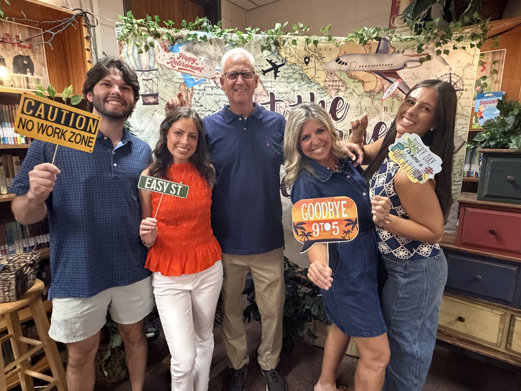 Five adults pose for a photo in a classroom. They hold signs and props. The background features a jungle theme with a map and plants.