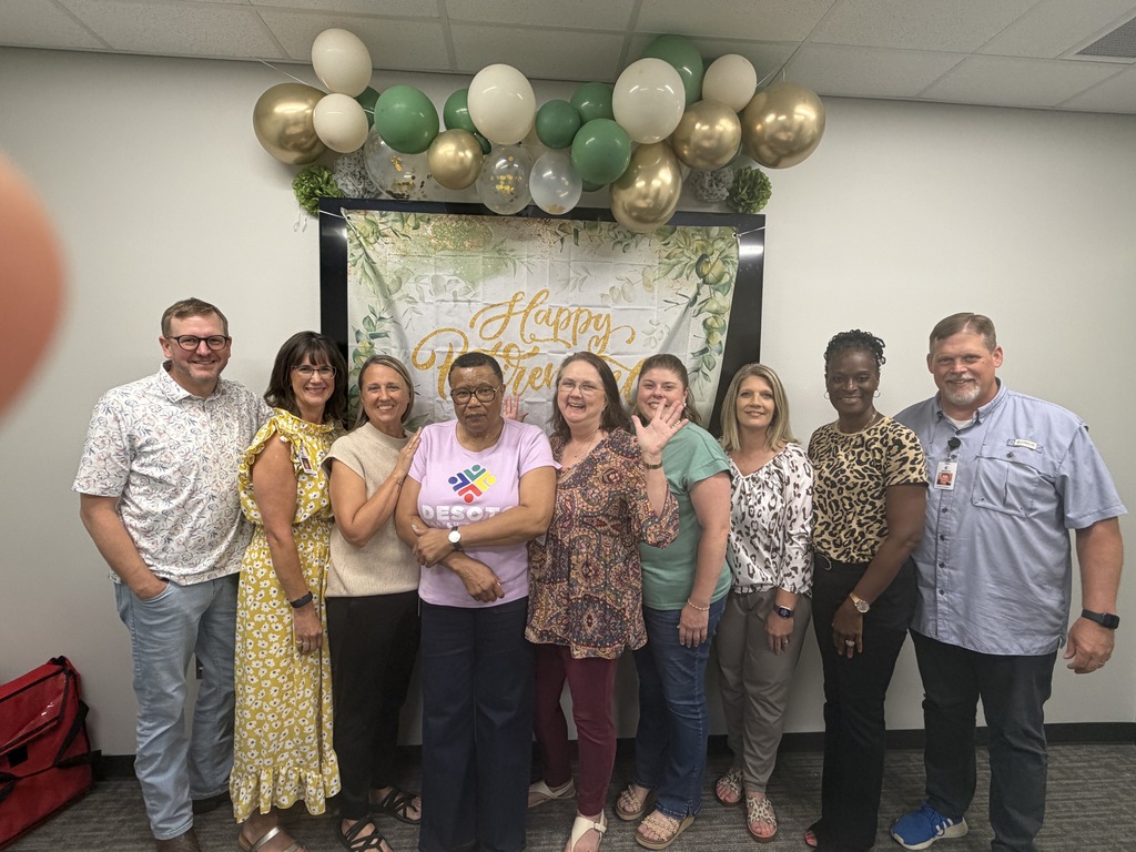 Nine people smile for a photo in front of a banner and balloons. The banner has "Happy" and "Year" written on it.