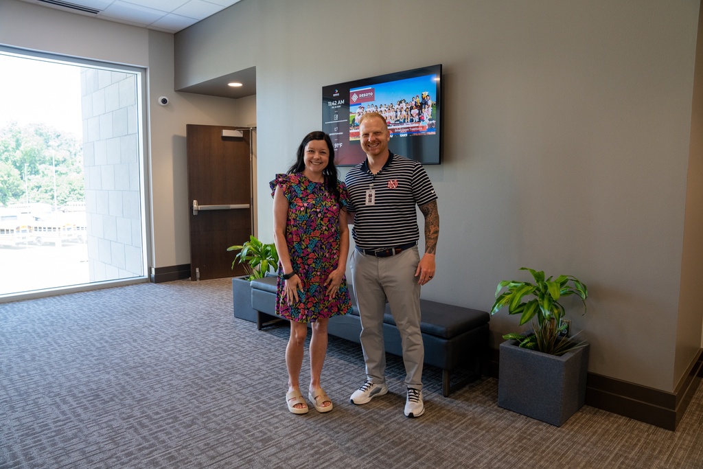 Two people stand in a room with a bench, plants, and a TV. The woman wears a floral dress and sneakers. The man wears a polo shirt, pants, and sneakers.