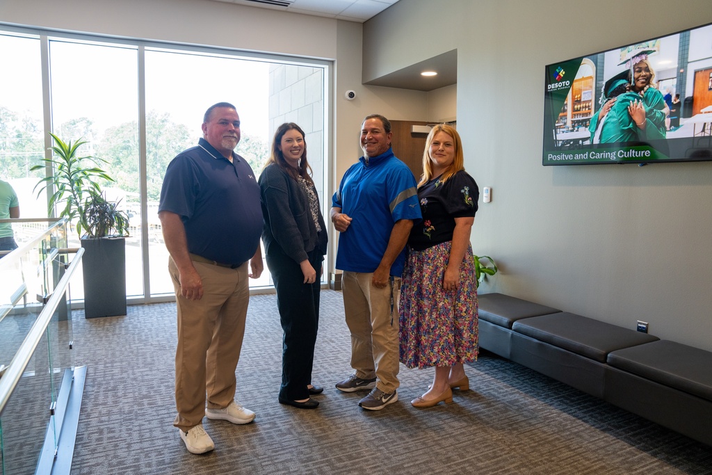 Five adults stand in a lobby area. A monitor is mounted on the wall, and a plant is on the floor.