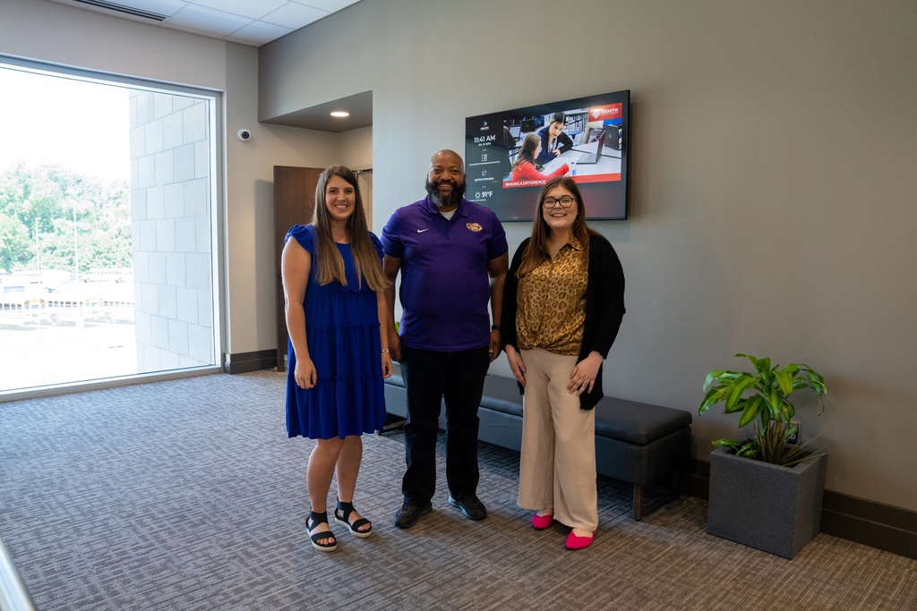 Three people stand in an office. A woman in a blue dress, a man in a purple shirt, and a woman in a black jacket. A TV is mounted on the wall.