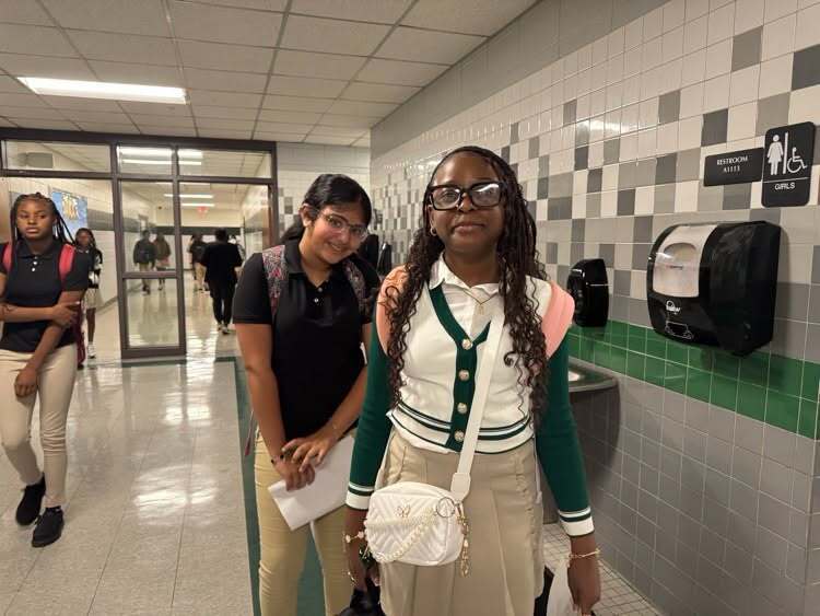 Three young women in a hallway, two holding papers, one with a white bag. Tiled walls and dispensers on one side.