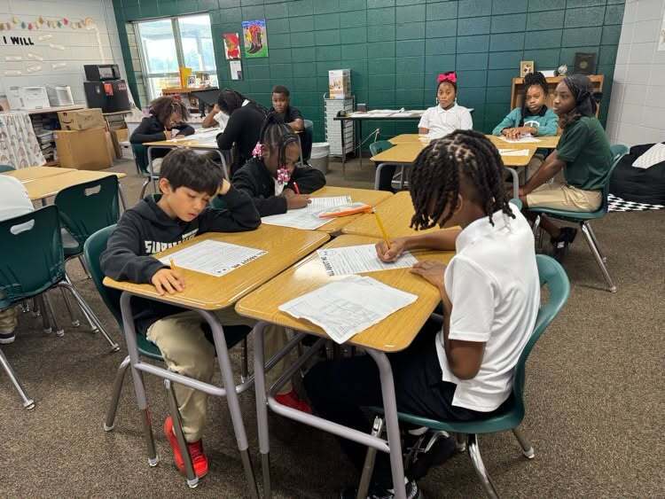 Students in a classroom focus on individual tasks. They sit at desks, with some using pens to write.