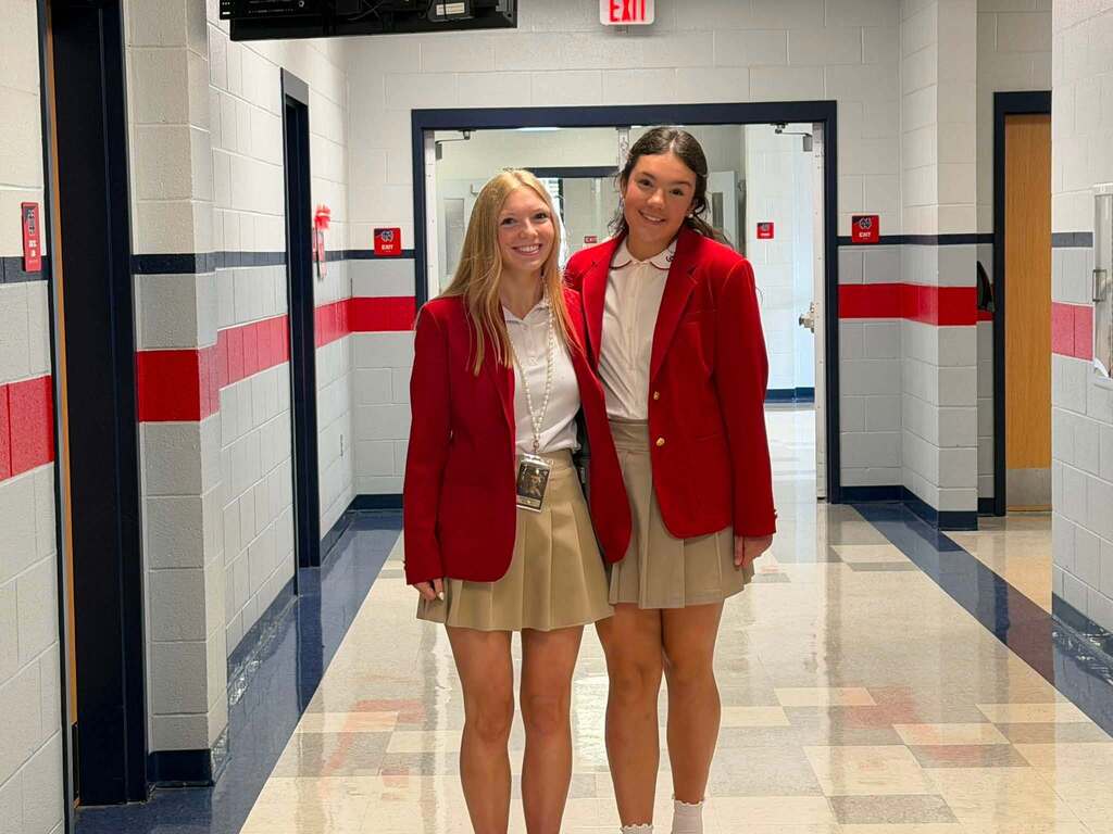 Two women stand in a hallway, both wearing red jackets and skirts, with white walls and red and white stripes.