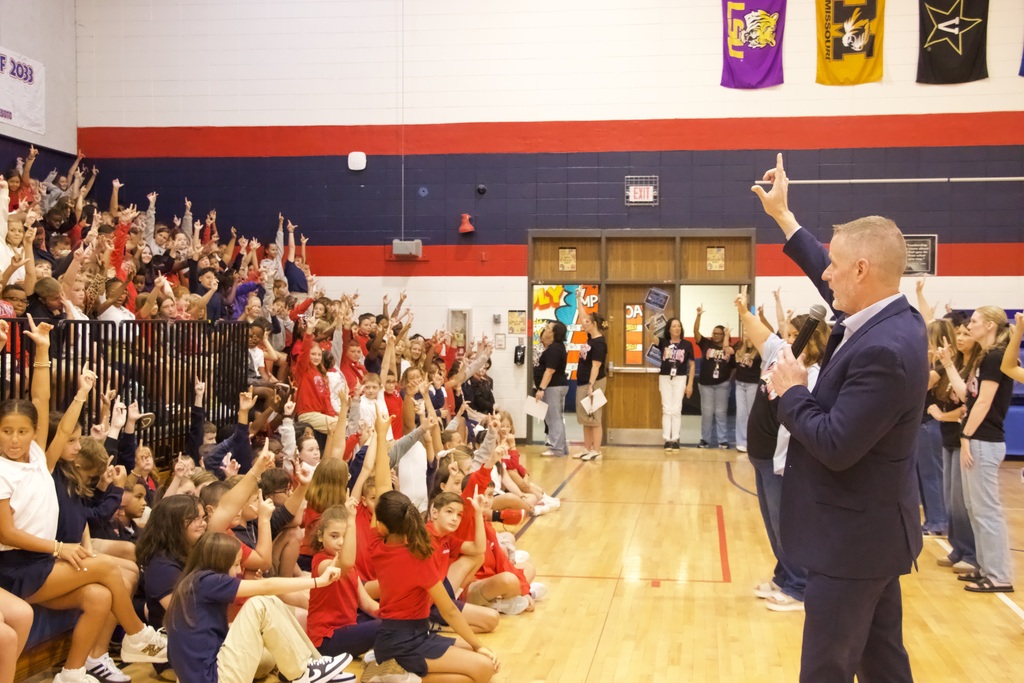 A man speaks into a microphone on a basketball court as a large audience of students watches.