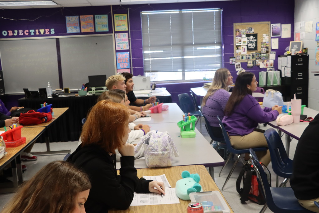 A classroom with purple walls. Students sit at tables, working with bags, pens, and books.