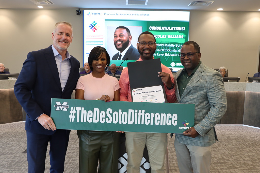 Four individuals stand, smiling, holding a certificate and a green sign reading "#TheDeSotoDifference" in front of a projector screen.