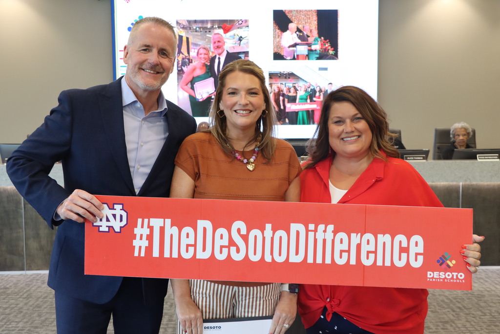 Three people stand behind a large sign that reads "#TheDeSotoDifference" in red letters, smiling and looking at the camera.