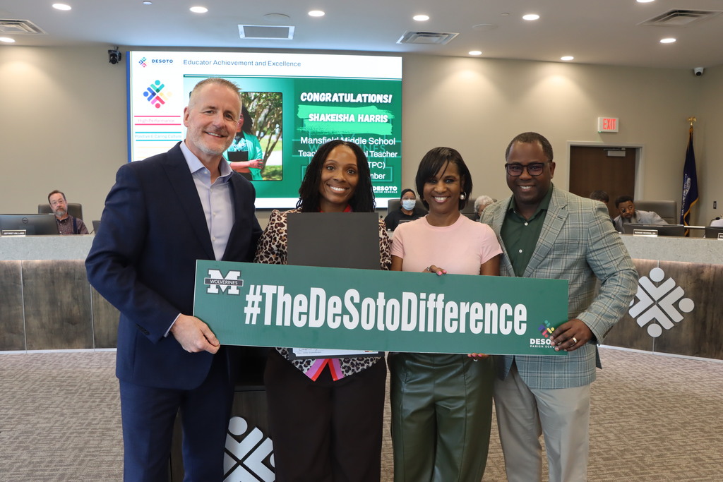 Four people pose with a green sign that reads "#TheDeSotoDifference." One holds a laptop, and a screen in the background displays text.