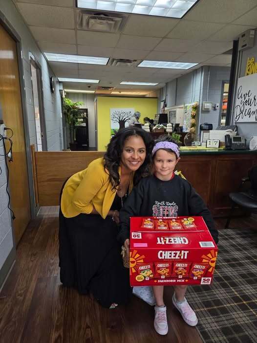 Woman in yellow and a child in black hold a large box of Cheez-Its. They sit on a floor.