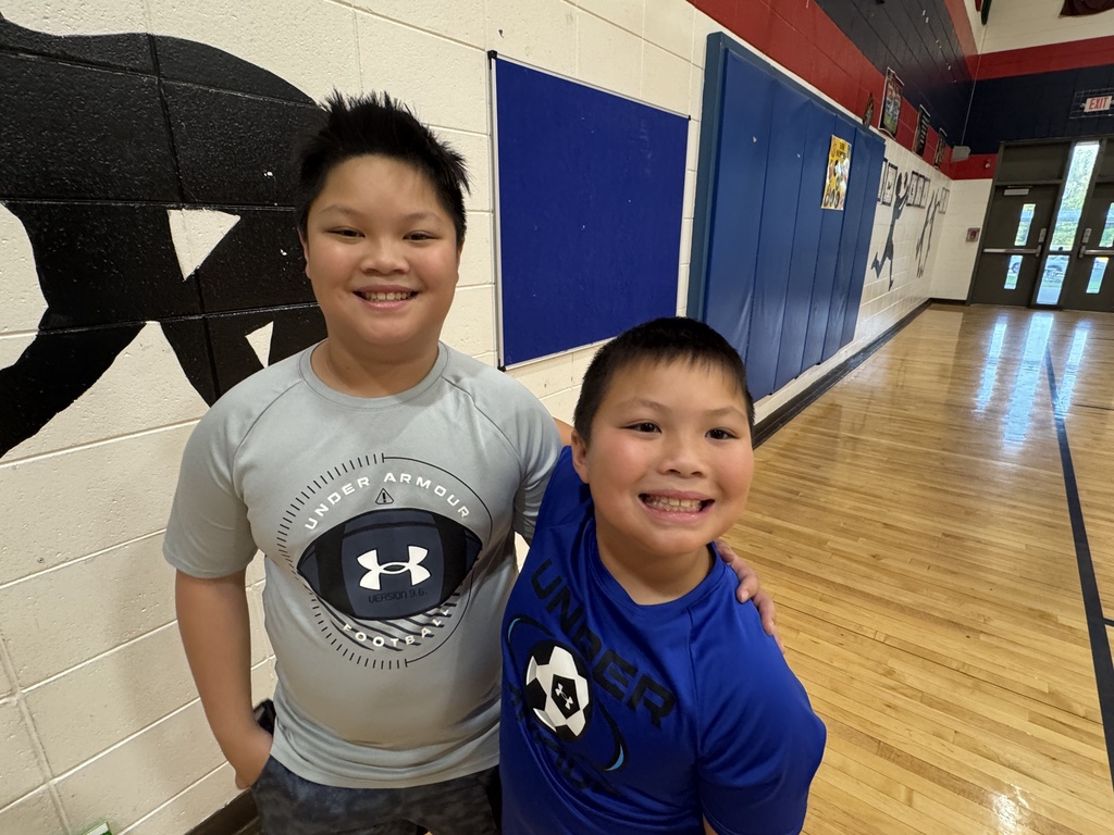Two children stand in a gym, smiling. One wears a gray Under Armour shirt, the other, a blue shirt.