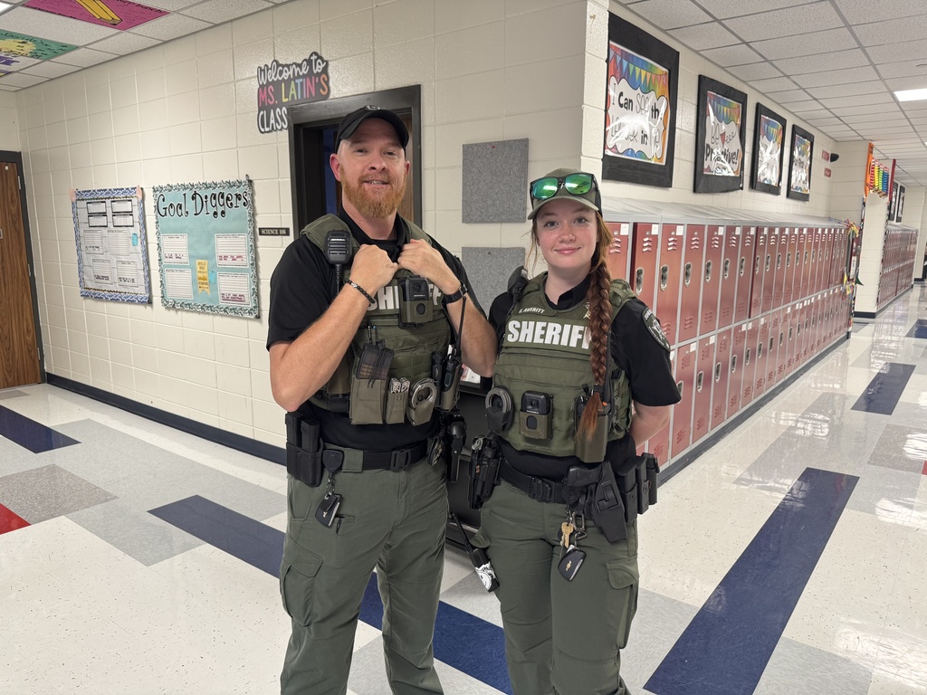 Two uniformed individuals in a school hallway, standing in front of lockers and bulletin boards.
