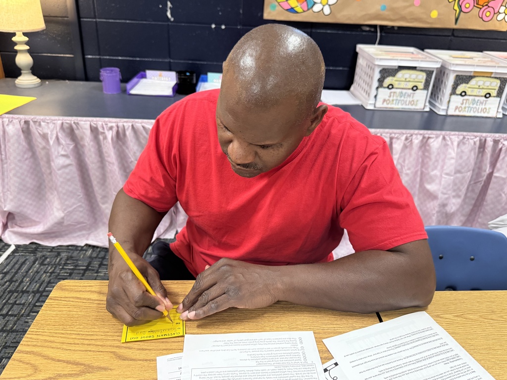 A man in a red shirt sits at a desk with papers and a yellow card, writing. A lamp and table with storage boxes are behind him.