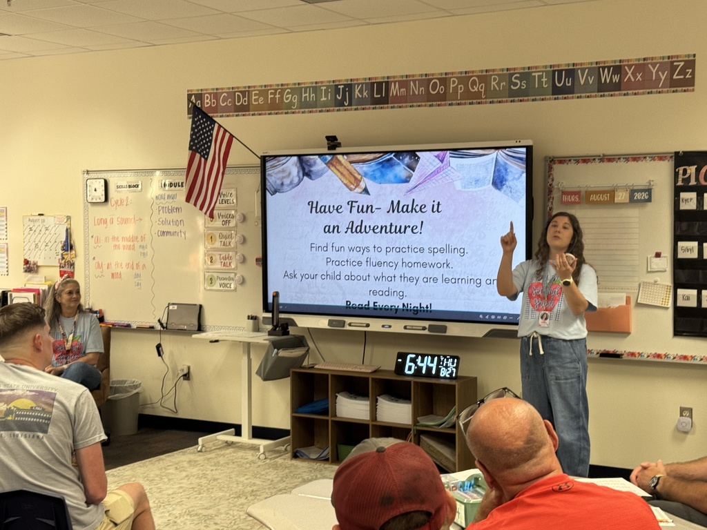 A teacher in a white t-shirt and pants stands in front of a screen giving a lesson.