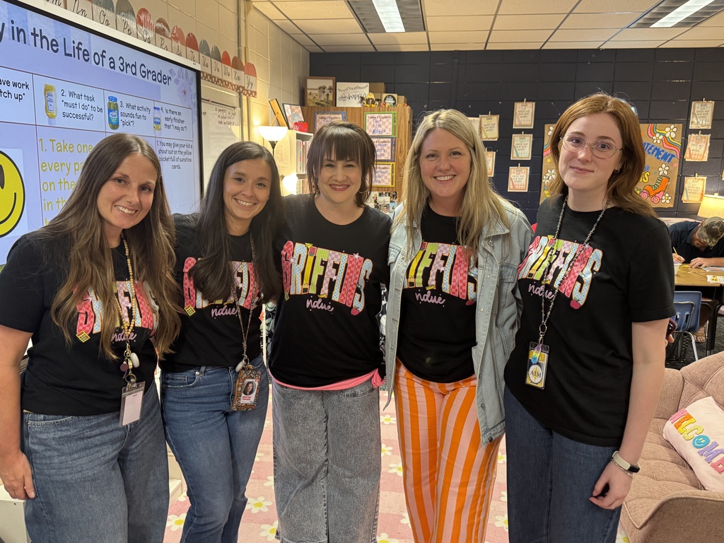 Five women stand in a room, all wearing black t-shirts with colorful designs. They stand in front of a white board with colorful drawings.