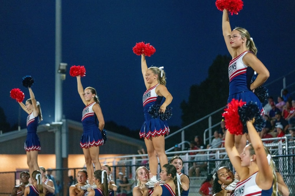 A group of cheerleaders, dressed in blue and red uniforms, performing a stunt in front of a stadium.