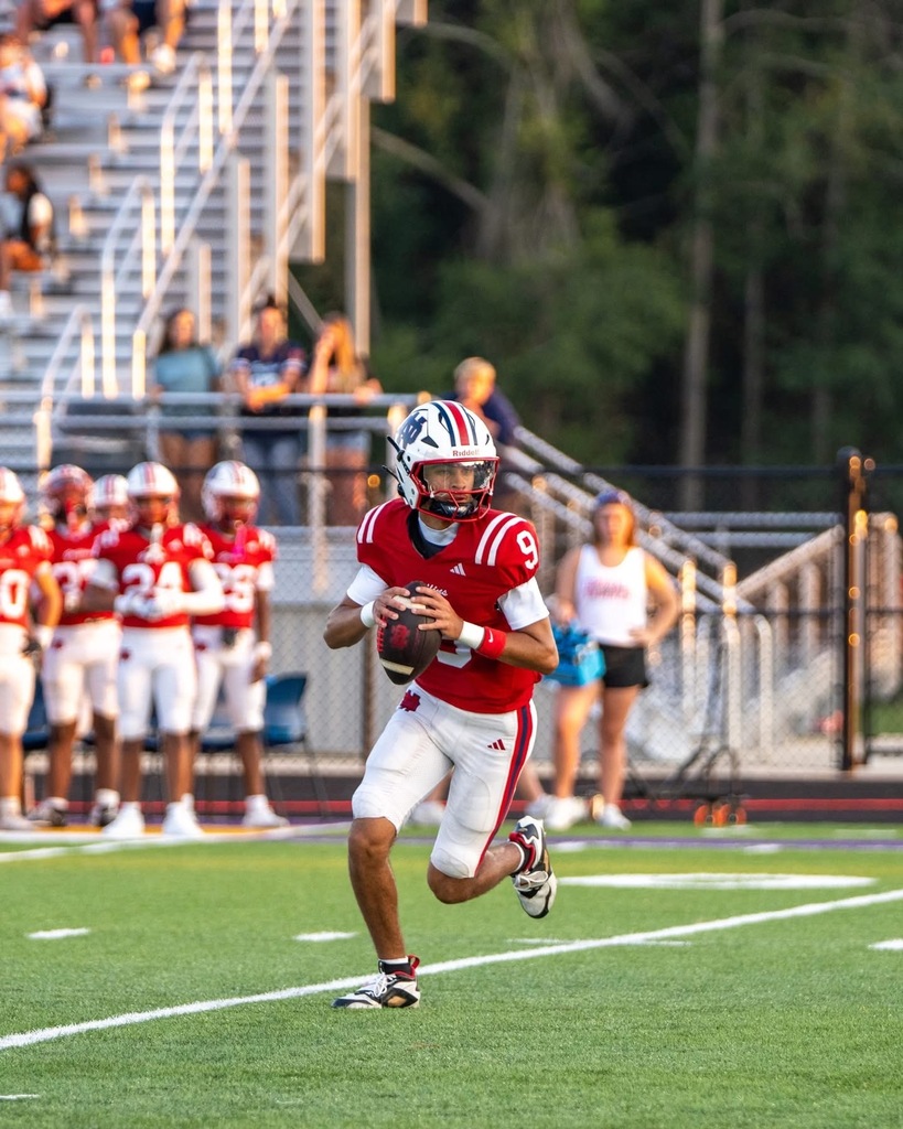 Football player in red jersey running with the ball. Players in white jerseys in background. White lines on the field.