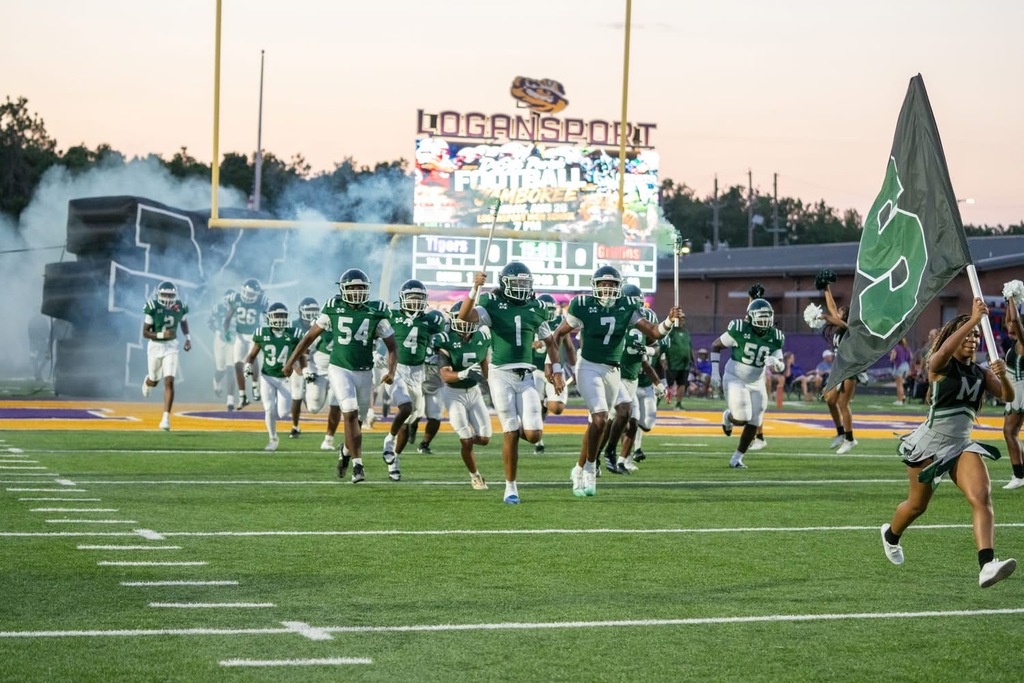 Football team on field, wearing helmets and jerseys. Smoke effect behind them. A person carries a flag.
