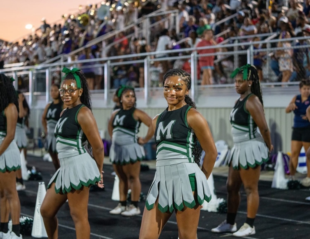 A group of cheerleaders in green uniforms perform on a field, with an audience seated in the stands.
