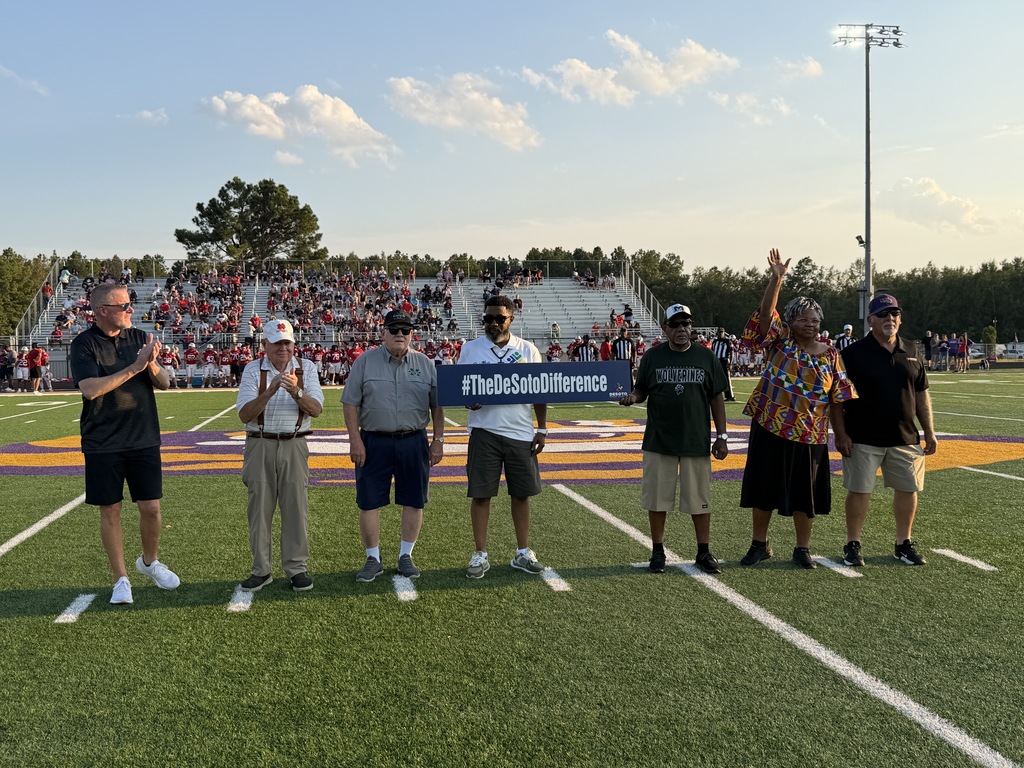 Six men on a football field holding a large banner. In the background, bleachers are full of people.