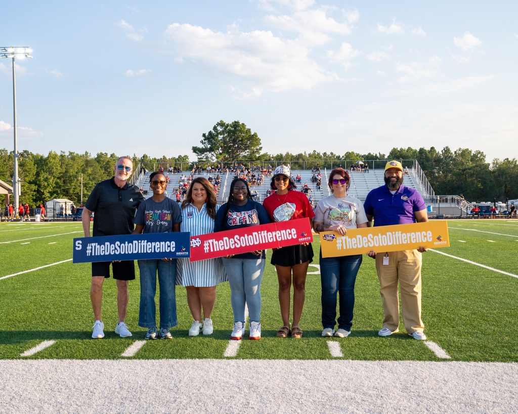 Group of people standing on a field holding signs that say "#TheDeSotoDifference" and "#WeMakeADifference".