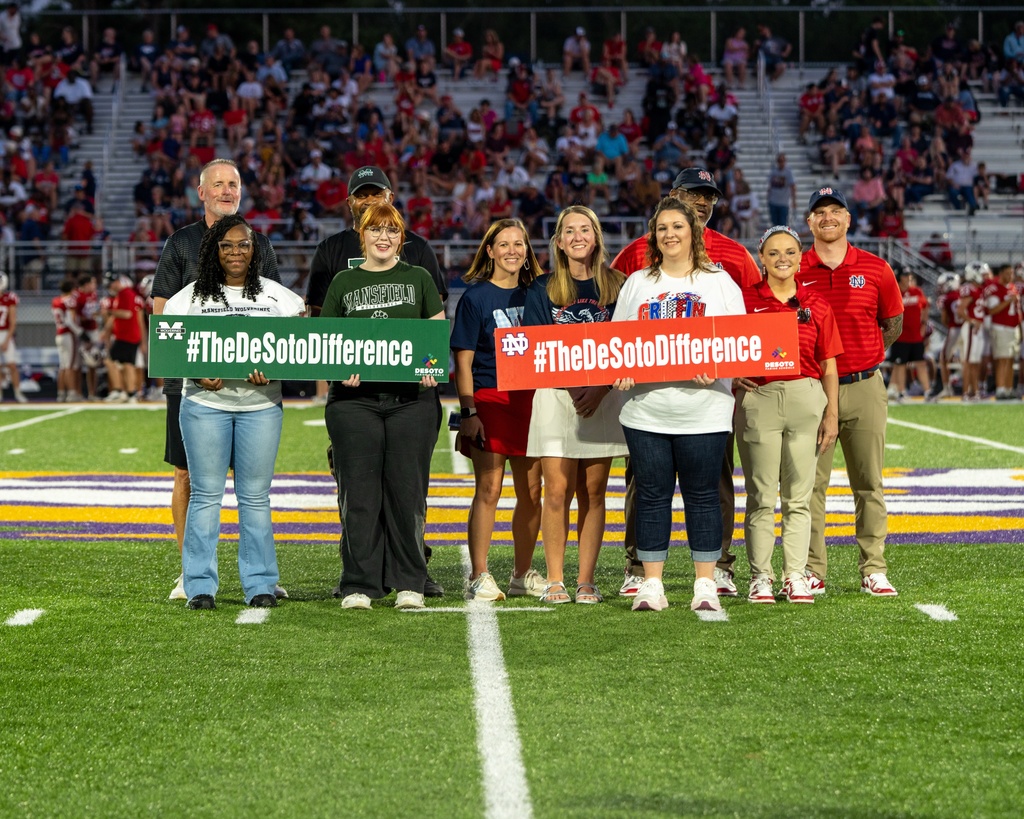 Group of people standing on a football field holding green signs with white writing. Audience in stands.
