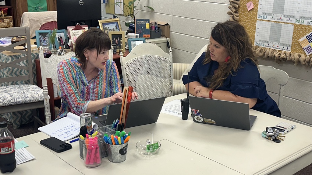 Two women seated at a table with laptops, papers, pens, pencils, and a bottle in front of them.