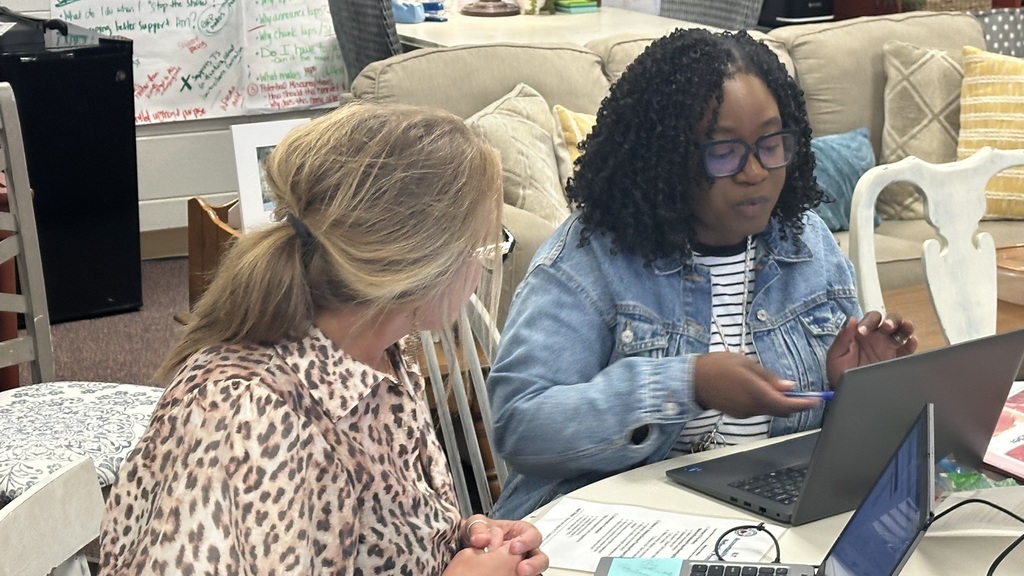 Two women in a room, one in a denim jacket, sit at a table with laptops.