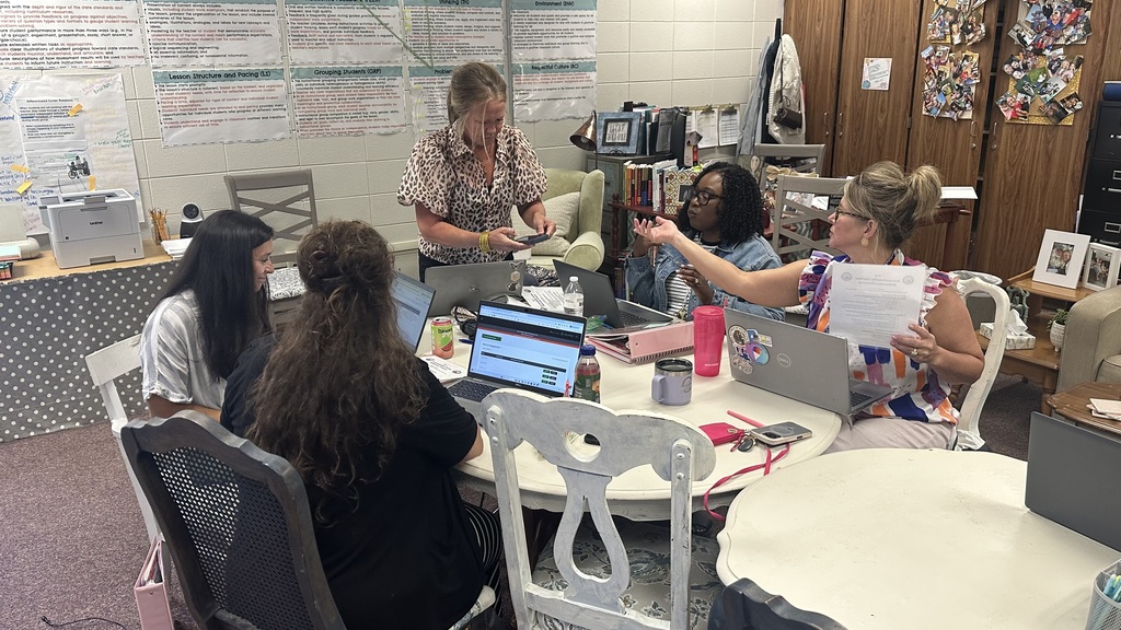 A group of women sits around a table with laptops and papers. A woman stands near a whiteboard.