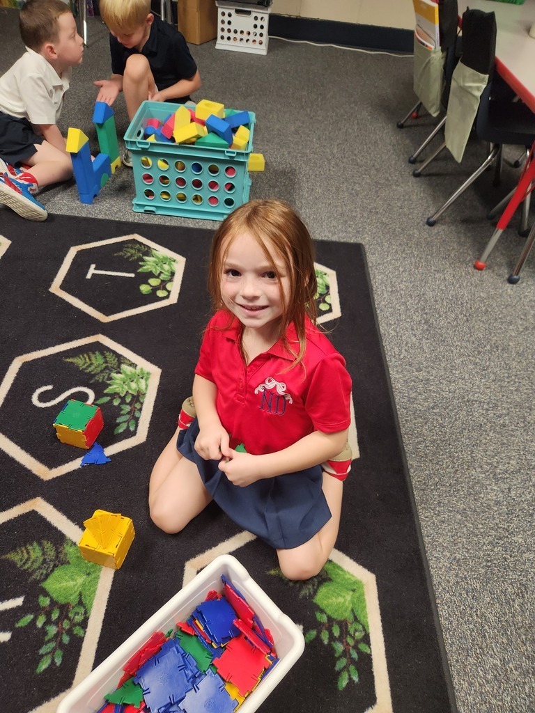 A girl sits on a floor, smiling. She holds a colorful block. Nearby, other children play with blocks.